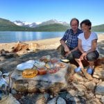 Kari and Håkon smiling and eating brunch outside on big rocks next the lake, Røssvatnet, in beautiful mountain surroundings.