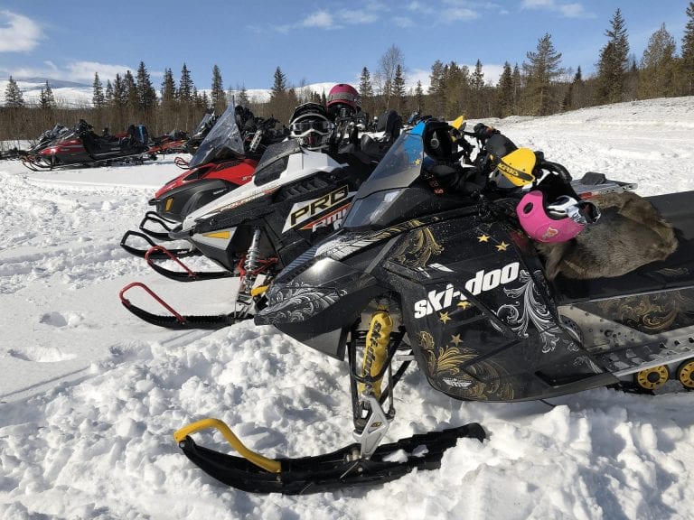 Picture of several colorful snowmobiles standing still in the snow on a sunny day.