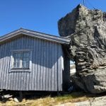 A small, grey cabin in the mountains during the summer with a huge rock “leaning” on the cabin.