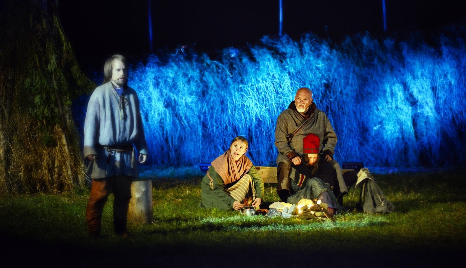 Main characters in old costumes sitting next to an open fire during local play, Klemetspelet, with blue lights behind.