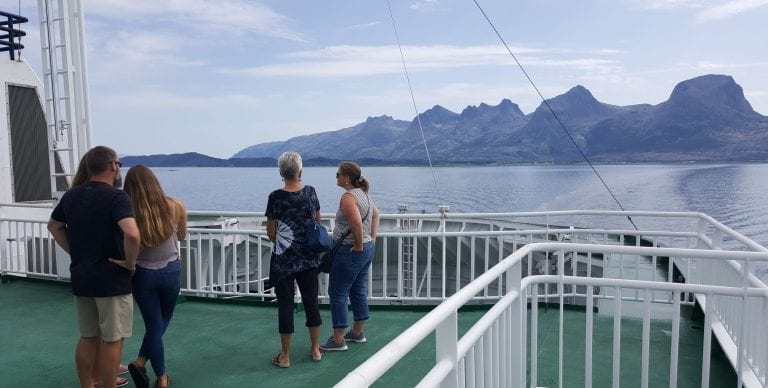 People standing outside on the top of the ferry on a summer day, with a great view of several grand mountains around them.