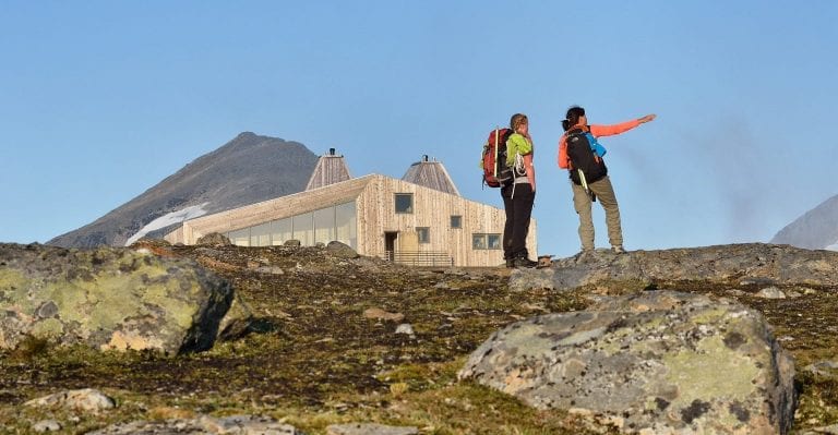 Two colorful hikers standing in front of the designer cabin, Rabothytta, surrounded by magnificent wilderness during summer.