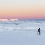 Skier surrounded by a white and open mountain landscape with a colorful, pink sunset in the horizon.