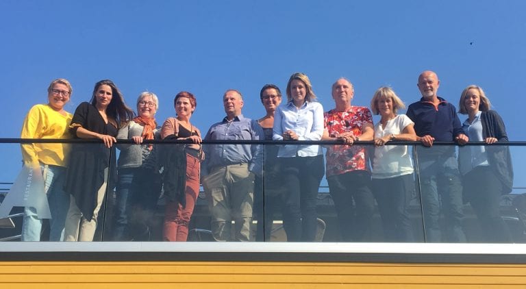 11 smiling partners of the Okstindan Park gathered standing up on a roof veranda in the sun.