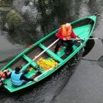 Duck race on the Røssåga river with two people sitting in a green boat trying to collect small floating ducks in the water.