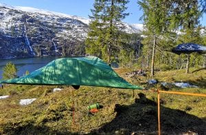 Tent hanging between trees above the ground, with a great view of the fjord and white mountains.