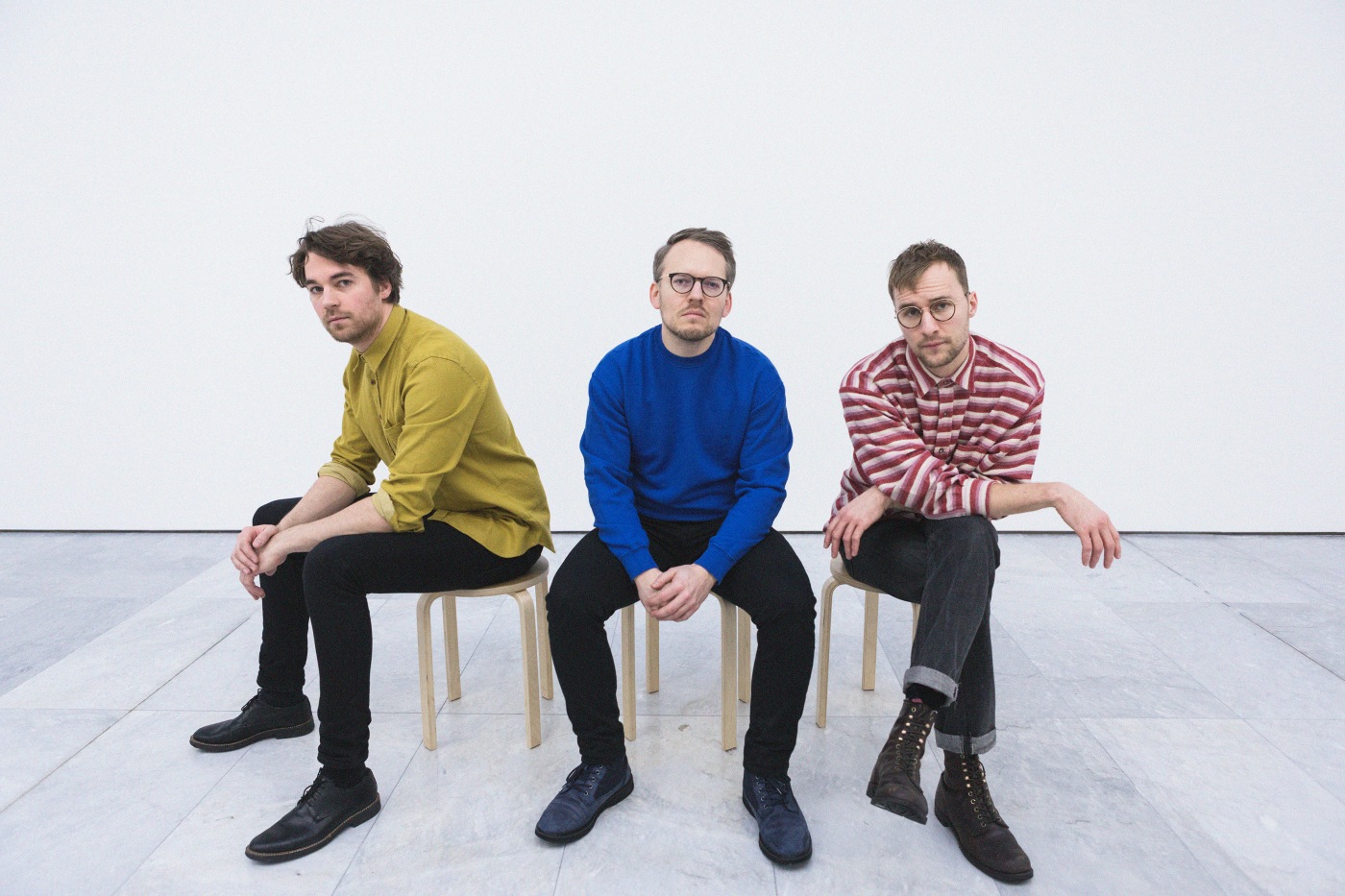 Three men dressed in colourful swethers sitting on wooden chairs in a white room.