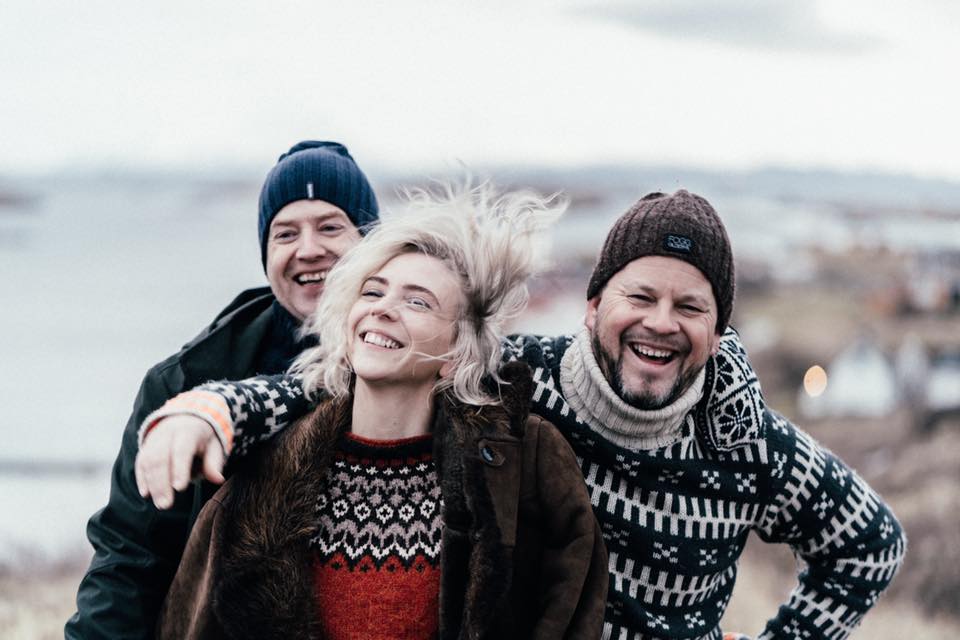 Tra hayppy musicians outside dressed in woolen sweaters and hats, two men and a woman.